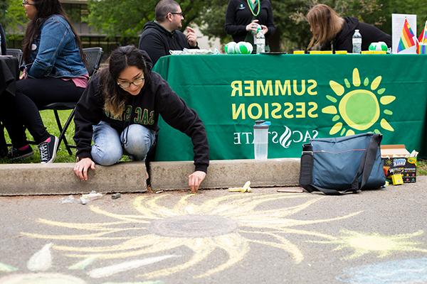 一张学生用粉笔在人行道上画画的照片, 桌子前挂着一条绿色横幅，上面写着“夏季会议”。